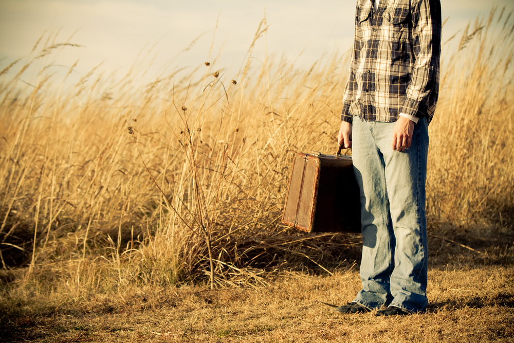 荒れた草原に呆然と立ち尽くす、古びたスーツケースを持った男性